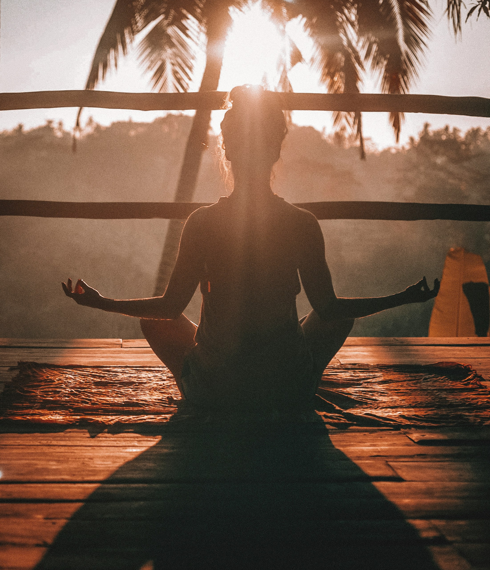 A woman meditating in silhouette on a wooden deck at sunset among palm trees
