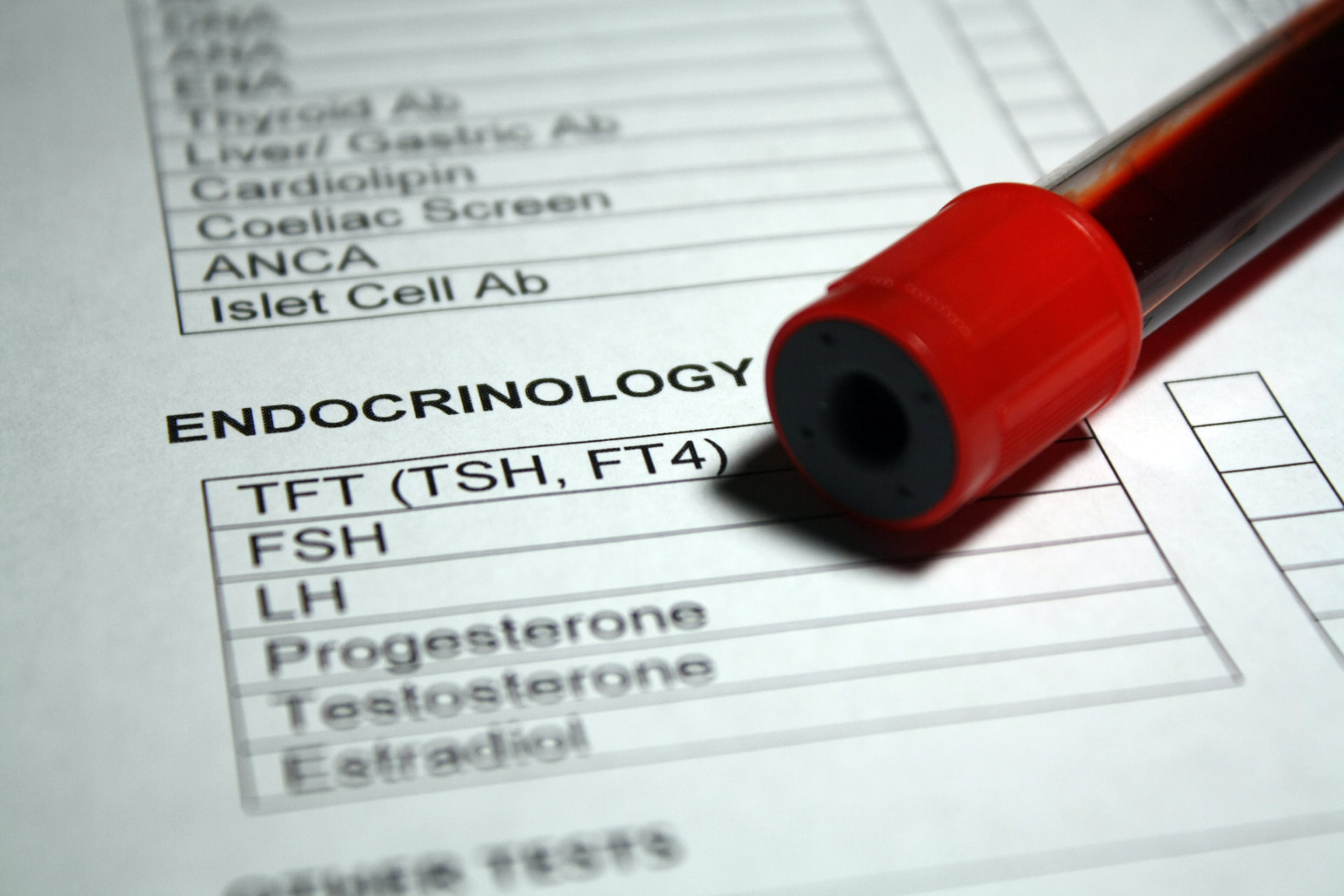 A red-capped blood collection tube resting on an endocrinology lab requisition listing TFT (TSH, FT4), FSH, LH, progesterone, testosterone, and estradiol