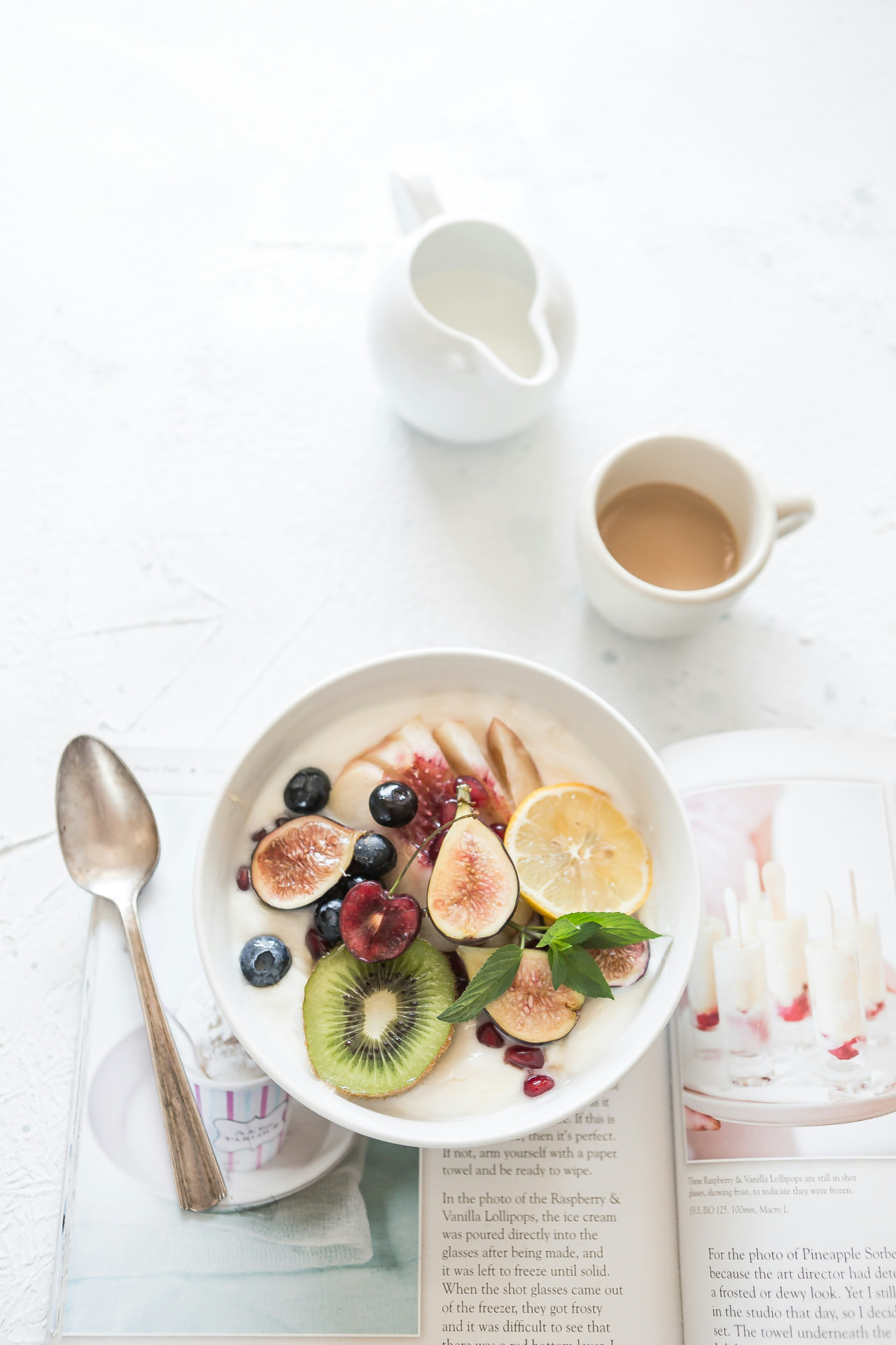 A breakfast bowl of yogurt, figs, kiwi, and berries beside a cup of coffee on a bright table