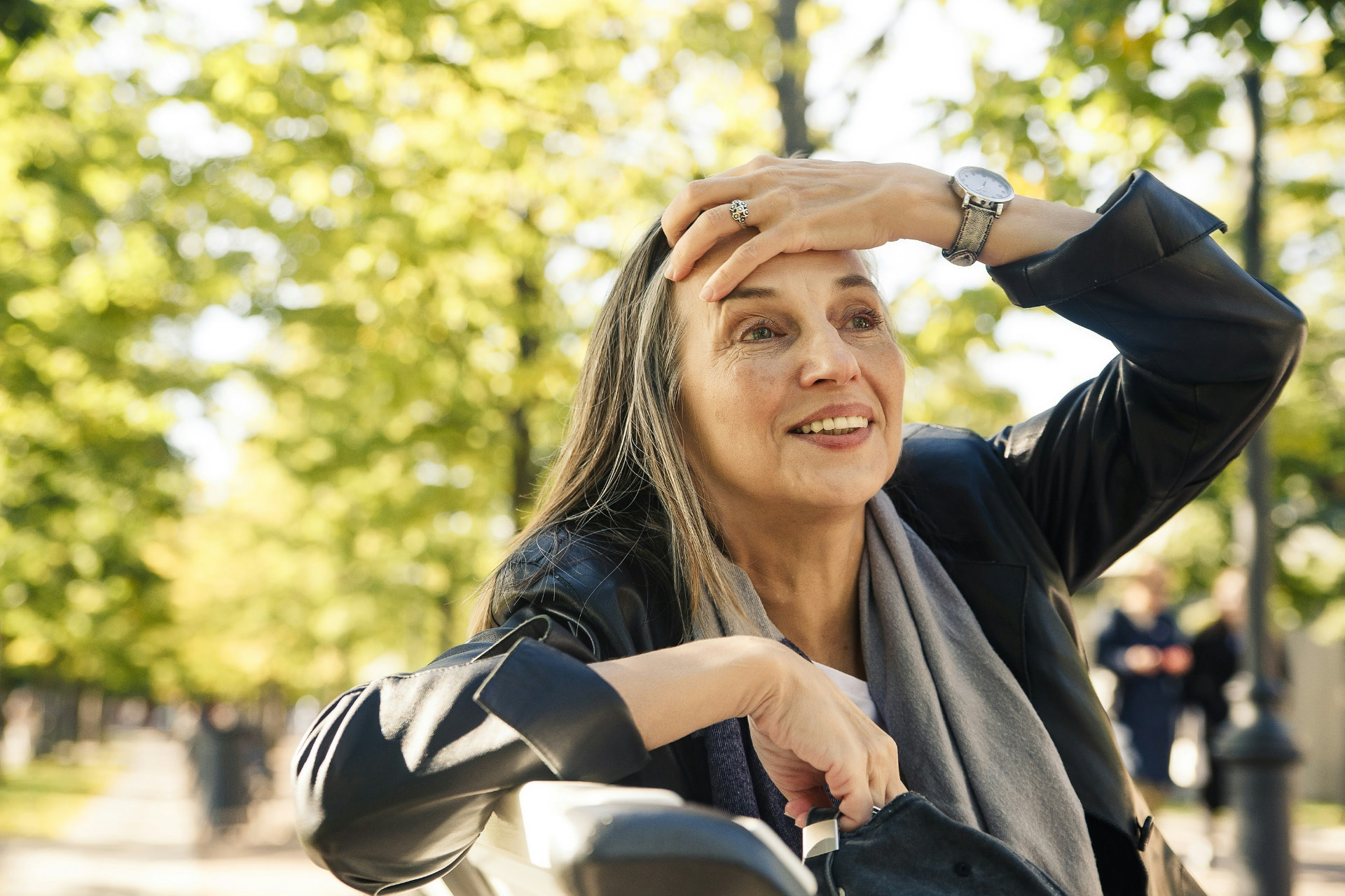 A smiling woman with long gray-streaked hair sits outdoors, shielding her eyes from the sun