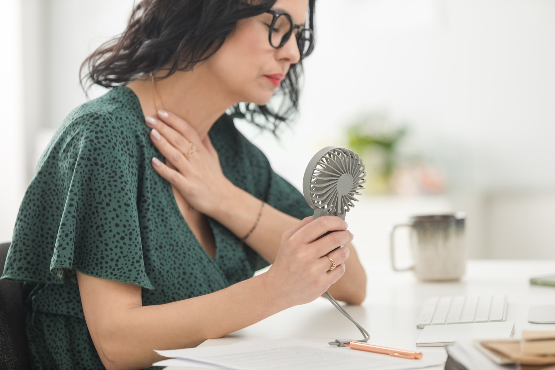 A woman at her desk holding a small handheld fan to her chest during a hot flash