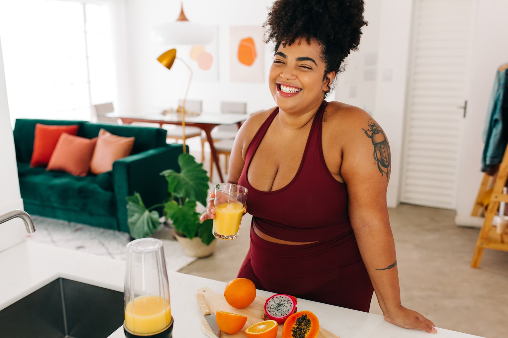 A woman in a bright home kitchen smiling while making a fresh fruit drink