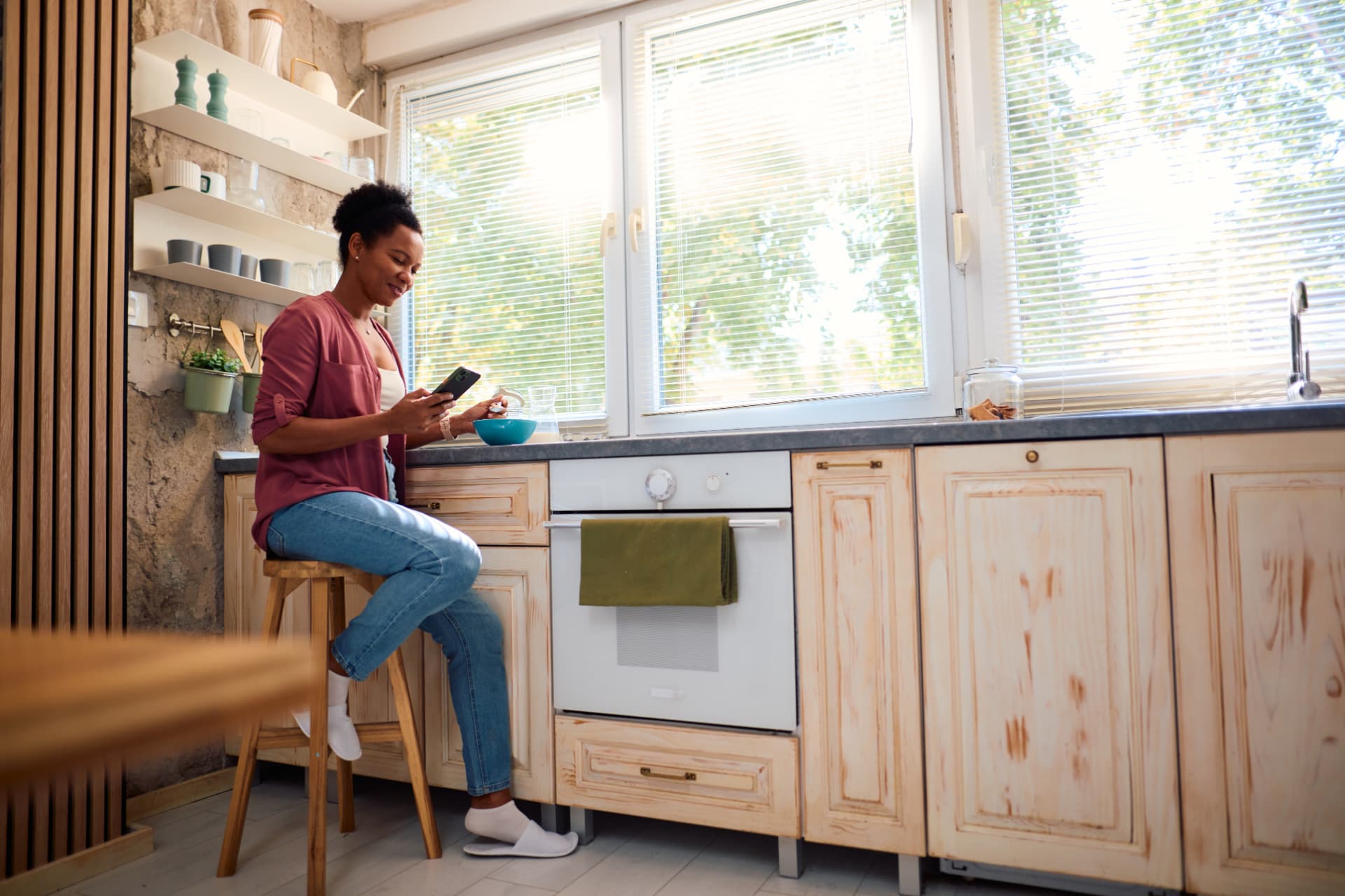 A woman sitting on a stool in a sunlit kitchen, resting with her phone and a small bowl