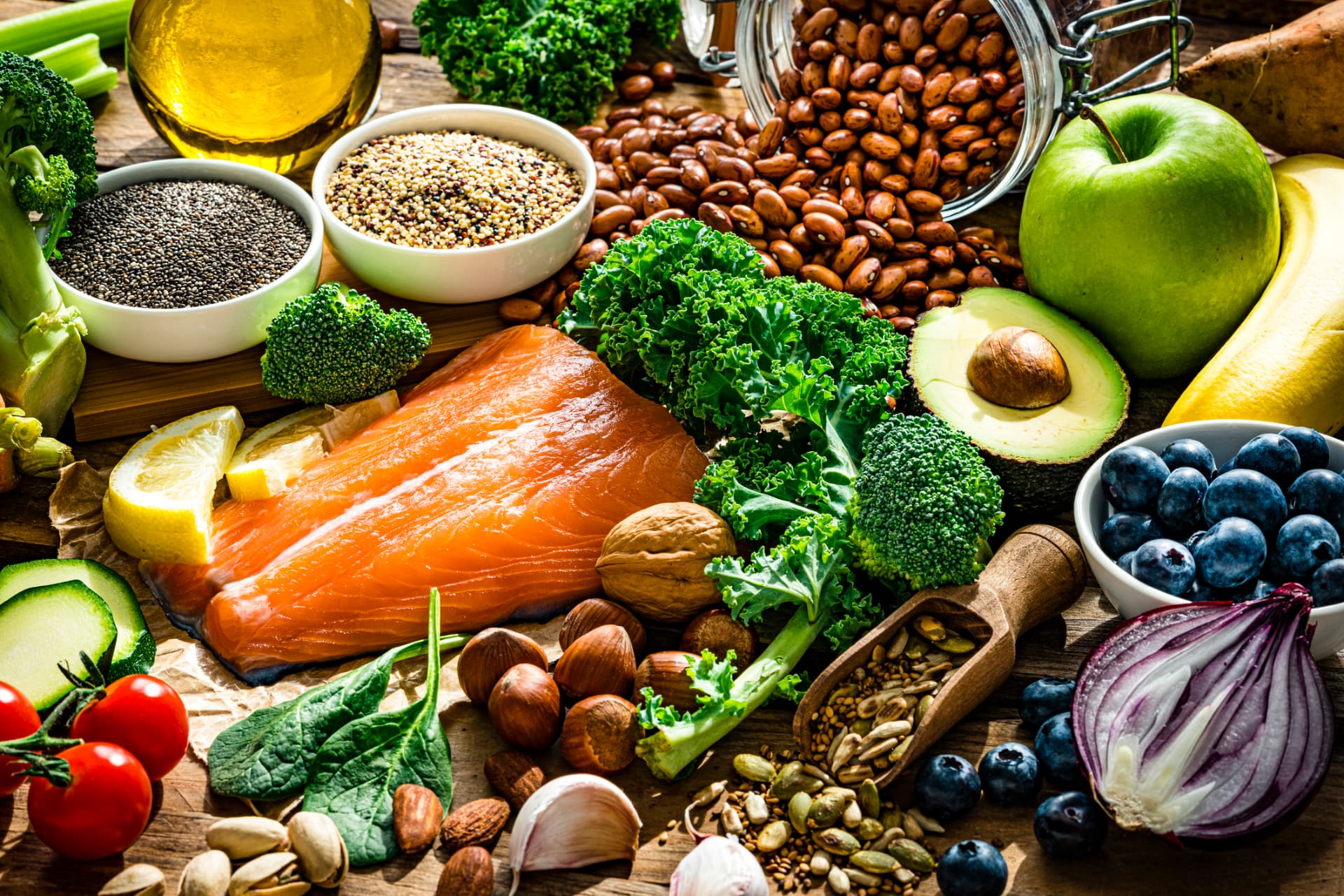 Wooden table spread with salmon, beans, quinoa, chia seeds, nuts, avocado, and fresh vegetables