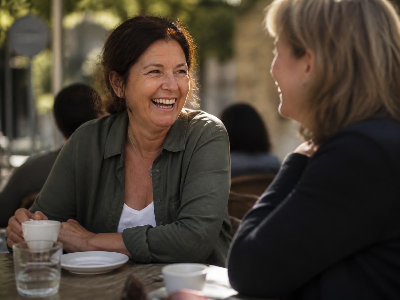 Two women in midlife laughing together over coffee at an outdoor cafe table